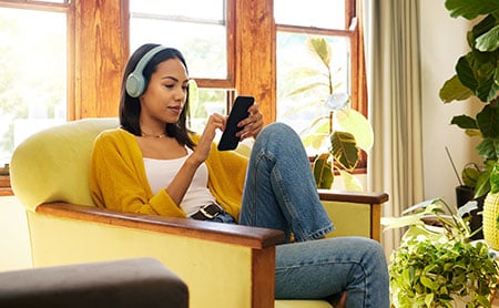 BIPOC woman wearing headphones sitting in a chair listening to a podcast on her phone