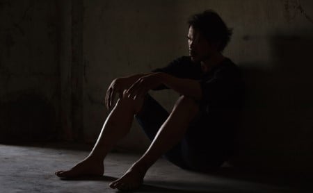 Depressed man sitting on the floor in a dark, concrete room.