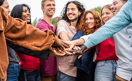 A diverse group of people in casual attire, symbolizing unity and teamwork by stacking their hands together in an outdoor setting.