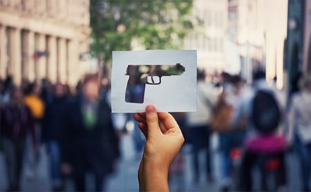 In focus in the foreground, a hand holds up a white paper square, the center of the square has the shape of a handgun cut out. An out-of-focus-crowd is in the background