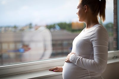 Pregnant woman looking out window with hand on her belly seeing her reflection