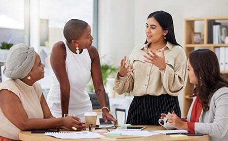 In an office setting, three women of color are focused on another woman of color standing and gesturing with her hands, possibly explaining or emphasizing a point.