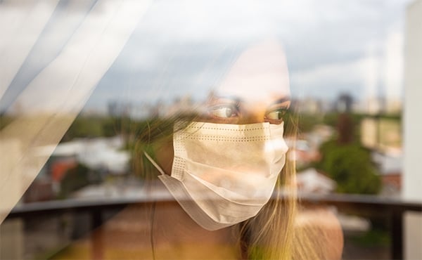 Woman wearing a protective face mask looks warily out of her window