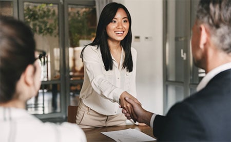 A person in a white shirt shaking hands with another person across a table while a third person observes.