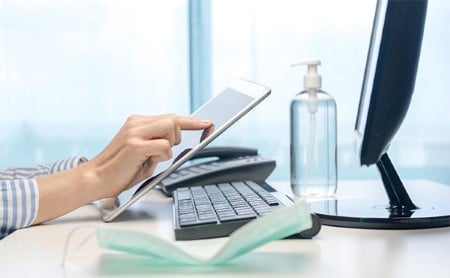 A woman's hands using a tablet while sitting at a desk with a disposable face mask and hand sanitizer on it.