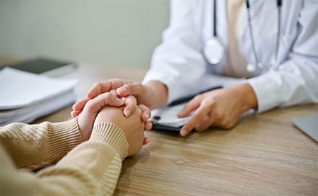 A doctor holding the hands of another person across a desk, suggesting a moment of comfort or reassurance