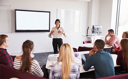 An instructor leading a session in a classroom with attendees facing a blank presentation screen.
