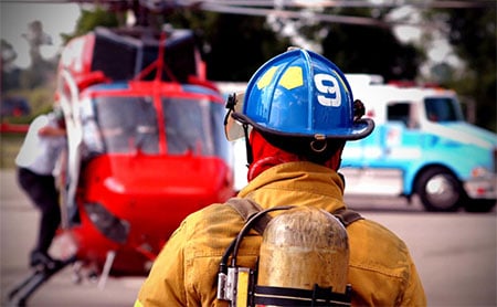 A firefighter in a helmet facing a red helicopter with an emergency vehicle in the background.