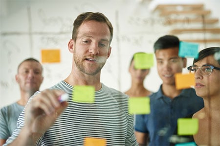 A group of individuals standing in front of a clear board with sticky notes, engaged in a collaborative activity.