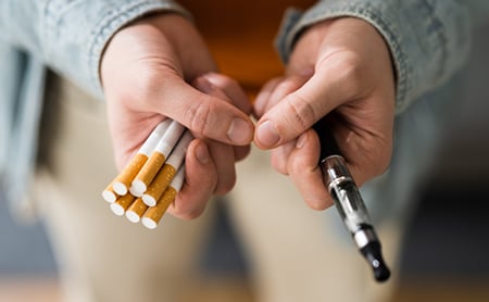 Hands holding combustible cigarettes in one hand and a single e-cigarette pen in the other