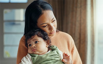 Woman of color in peach sweater resting her right cheek on a curly-haired, chubby cheeked baby in a green jumper looking off to the left