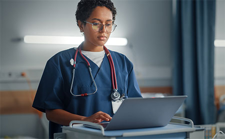 A healthcare professional in blue scrubs with a stethoscope around the neck is using a laptop on a mobile desk.
