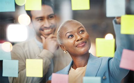 A woman of color is arranging a vibrant array of sticky notes on a glass wall, indicating a brainstorming or planning session, as her colleague observes the process with a smile from the background.