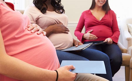 Mid-section shot of three pregnant individuals with hands on their bellies sitting in a waiting room.