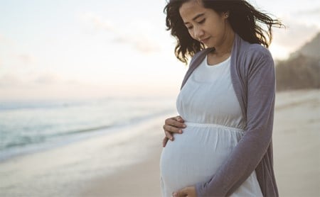 A pregnant woman stands on the beach cradling her stomach