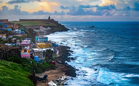 A picturesque view of a coastal scene in Puerto Rico, featuring multicolored houses along the shore, waves crashing against the rocky coast, and a distant lighthouse under an overcast sky.
