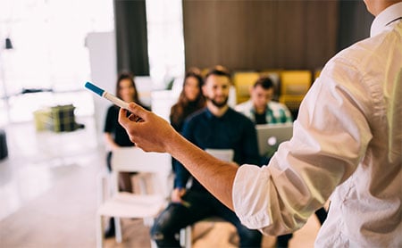 Person holding a marker in front of a seated group in a classroom.