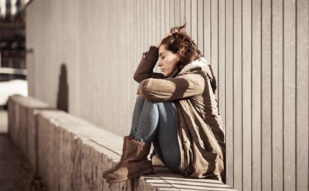 A woman sits against a metal fence looking sad and desperate, her knees brought up to her chest and her head resting in her hand
